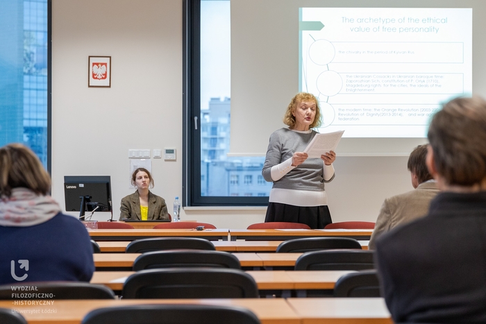 Kobieta przedstawiająca referat, wnętrze auli, w tle ekran z prezentacją w j. angielskim