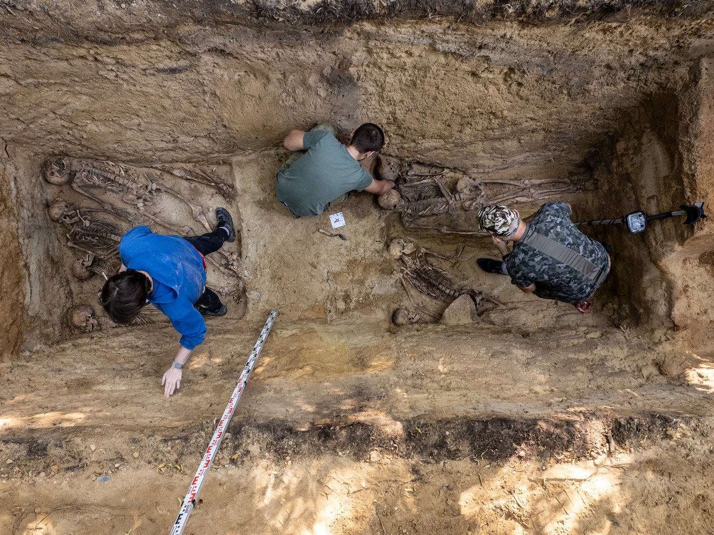 Widoczny wykop jamy grobowej. Na dnie ludzkie szkielety. Nad nimi pracujący archeolodzy.
